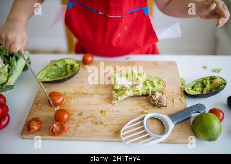 Weibliche Kinderhände verteilen Avocado auf einer Brotscheibe über dem Holzschneidebrett. Yung Mädchen am weißen Tisch schneiden Gemüse. Kinder bereiten Essen zu Stockfoto