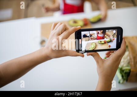 Weibliche Hände mit Smartphone Aufnahme liebenswert kleines Mädchen Verbreitung Avocado auf Scheibe Brot. Niedliches kleines Kind schneidet Gemüse auf Schneidebrett. Stockfoto