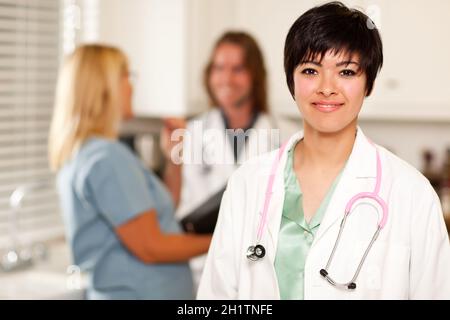 Der hübsche Latino-Arzt schlint an der Kamera, während Kollegen hinter ihr reden. Stockfoto