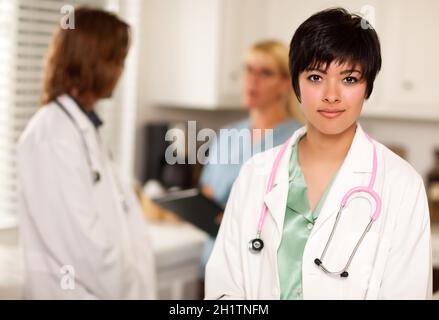 Der hübsche Latino-Arzt schlint an der Kamera, während Kollegen hinter ihr reden. Stockfoto