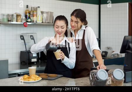 Junger asiatischer Barista mit einem Lächeln gießt Milch aus dem Krug in eine Papierkaffeetasse. Kaukasische Assistentin in einer Schürze steht neben ihr und beobachtet sie. Mornin Stockfoto