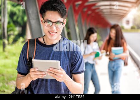 Glücklich junge asiatische Mann Student Lesen auf Tablet-Computern auf dem Campus. Es gibt Gruppen von Freundinnen dahinter, Bildung zurück zur Universität Konzept Stockfoto