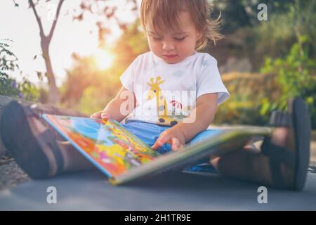 Entzückender kleiner Junge mit einem Buch sitzt im Garten in einem schönen sonnigen Tag. Genießt es, sich die bunten Bilder auf den Seiten des Buches anzuschauen. Stockfoto