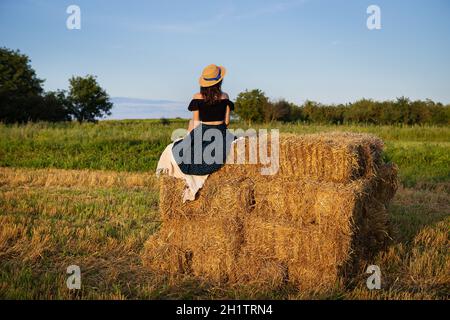 Junge schöne Frau in einem Strohhut schaut auf die untergehende Sonne, das Mädchen sitzt auf einem großen Haufen Strohballen. Stockfoto