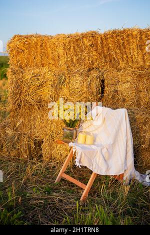 Picknick auf dem Feld in der Nähe von Strohballen. Die untergehende Sonne. Rustikaler Stil - Holzstuhl, Karo, Blumenstrauß, Kerzen Stockfoto