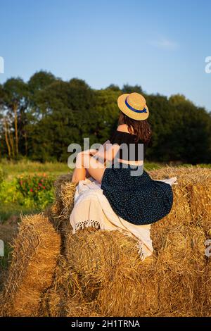 Junge schöne Frau in einem Strohhut schaut auf die untergehende Sonne, das Mädchen sitzt auf einem großen Haufen Strohballen. Warmer Sommerabend Stockfoto