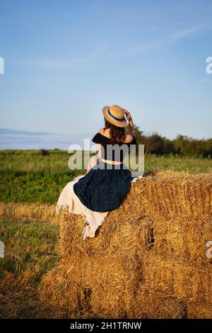 Junge schöne Frau in einem Strohhut schaut auf die untergehende Sonne, das Mädchen sitzt auf einem großen Haufen Strohballen. Warmer Sommerabend Stockfoto