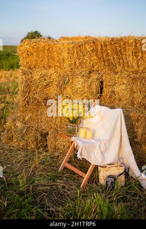 Picknick auf dem Feld in der Nähe von Strohballen. Die untergehende Sonne. Rustikaler Stil - Holzstuhl, Karo, Blumenstrauß, Kerzen Stockfoto