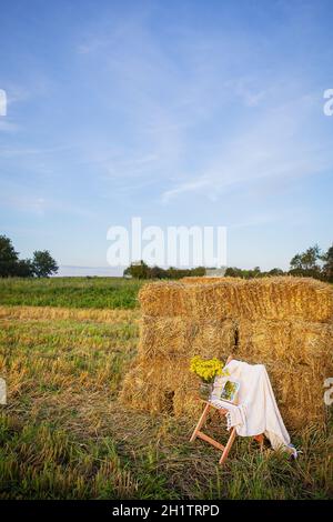 Picknick auf dem Feld in der Nähe von Strohballen. Die untergehende Sonne. Rustikaler Stil - Holzstuhl, Karo, Blumenstrauß, Spiegel. Romantisches Date Stockfoto