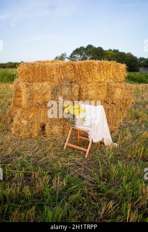 Picknick auf dem Feld in der Nähe von Strohballen. Die untergehende Sonne. Rustikaler Stil - Holzstuhl, Karo, Blumenstrauß, Spiegel. Romantisches Date Stockfoto