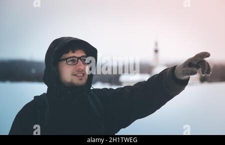 Ein junger lächelnder Mann in einer schwarzen Jacke mit Kapuze zeigt irgendwo hin, im Winter draußen Stockfoto