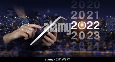 Ladejahr 2021 bis 2022. Konzept starten. Hand Touch weißes Tablet mit digitalem Hologramm 2022 Zeichen auf Stadt dunkel verschwommen Hintergrund. Willkommen im Jahr 2022. Stockfoto