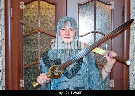 Ein Mann, gekleidet in ein ritterliches Kettenhemd zu Hause in seinem Zimmer. Knight's Schwert in den Händen eines Mannes. Ritterliche Kette Rüstung und Schwert. Einbau eines knightl Stockfoto