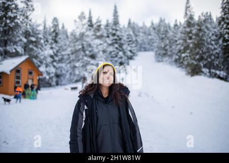 Glückliche Frau, die mit einem wunderschönen verschneiten Wald und einer kleinen Hütte als Hintergrund steht. Lächelnde Frau, umgeben von verschneiten Pinien. Abenteuerlicher Winterurlaub Stockfoto