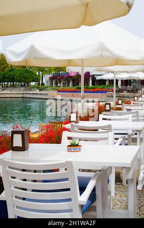 Weiße Tische und Stühle auf der Terrasse eines gemütlichen Sommercafés mit Blick auf das Wasser in Turgutreis Stadt, Türkei. Stockfoto