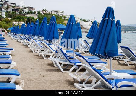 Reihen von leeren blauen Sonnenliegen und Sonnenschirmen am Strand. Windiger Sommertag. Camel Beach an bewölktem Tag, Türkei. Stockfoto