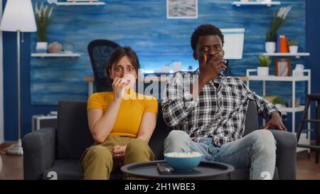 POV von schockierten interracial paar schaurig Film im Fernsehen zu Hause. Multiethnische Ehemann und Ehefrau haben Angst und verdecken die Augen, während sie die Kamera im Wohnzimmer betrachten Stockfoto