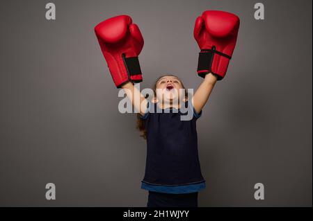 Konzept der Schlag auf die Preise auf grauem Hintergrund mit Kopierraum. Die kleine, hübsche Boxerin mit roten Boxhandschuhen hebt die Hände in der Siegerpositiono Stockfoto