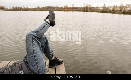 Ein Mädchen in blauen Jeans sitzt am See. Eine junge Frau ruht am Fluss und sitzt am Rande einer Holzbrücke. Zwei Beine, die sich an einem See entspannen Stockfoto