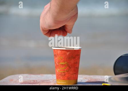 Kaffeepause an der Küste. Der Mann fügt etwas Zucker in die Einweg-Kaffeetasse aus Kunststoff Stockfoto