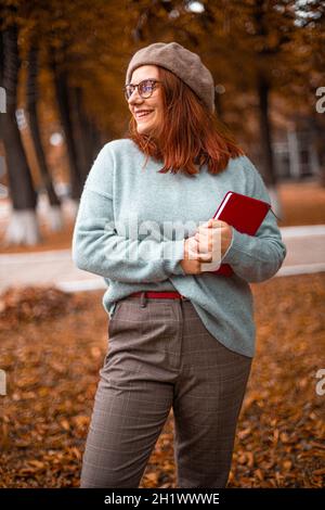 Junge emotionale fröhlich Füchshaar Frau in Brillen und Herbstkleidung und hält Notizbücher in der Hand, während sie wegschaut Stockfoto