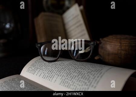 Alte getragene Gläser, die auf einem geöffneten Buch mit Büchern und Uhr im Hintergrund ruhen. Lesezeitkonzept Stockfoto