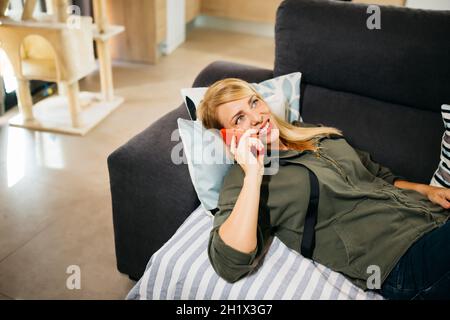 Lächelnde blonde Frau mittleren Alters, die auf einem Sofa liegt und in einem Wohnzimmer telefoniert Stockfoto