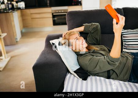 Lächelnde blonde Frau mittleren Alters, die auf einem Sofa liegt und einen Videoanruf auf einem roten Smartphone in einem Wohnzimmer führt Stockfoto