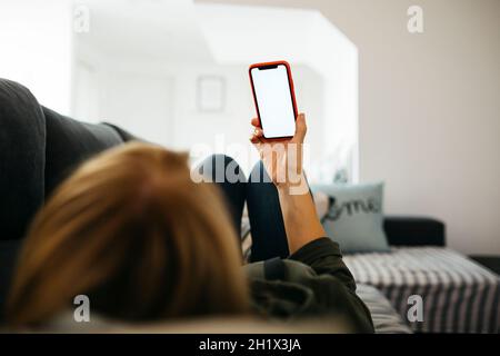 Hand einer lächelnden blonden Frau mittleren Alters, die ein rotes Smartphone in der Hand hält, während sie auf einem Sofa in einem Wohnzimmer liegt Stockfoto