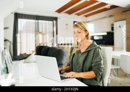 Lächelnde blonde Frau im mittleren Alter, die einen Laptop in einem Wohnzimmer benutzt Stockfoto