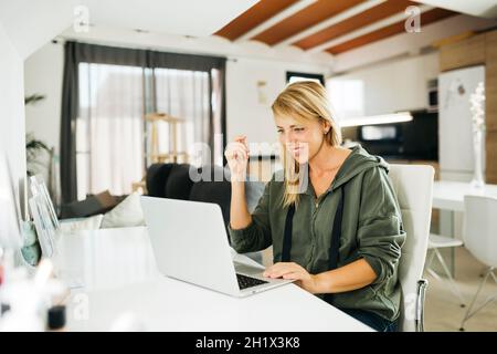 Lächelnde blonde Frau im mittleren Alter, die einen Laptop benutzt und einen Videoanruf in einem Wohnzimmer führt Stockfoto