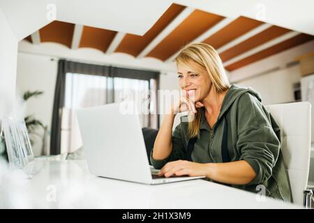 Lächelnde blonde Frau im mittleren Alter, die einen Laptop benutzt und einen Videoanruf in einem Wohnzimmer führt Stockfoto