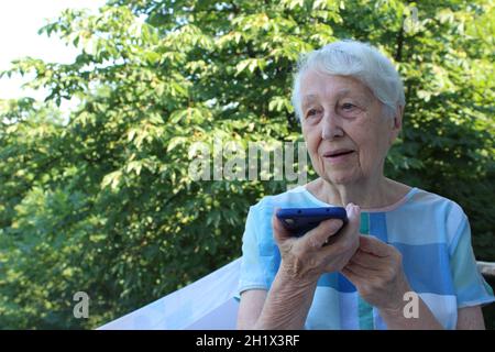 Charmante ältere Frau in rechteckiger, schwarz umrandter Brille mit Spracherkennungsfunktion auf dem Handy. Moderne ältere weibliche Holding elektronische gad Stockfoto