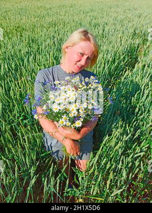 Porträt einer Frau mit Blumenstrauß von Kamillen und Kornblumen im Weizenfeld. Porträt einer ländlichen Frau mit Blumen. Frau mit Blumenstrauß aus weißem Feld da Stockfoto