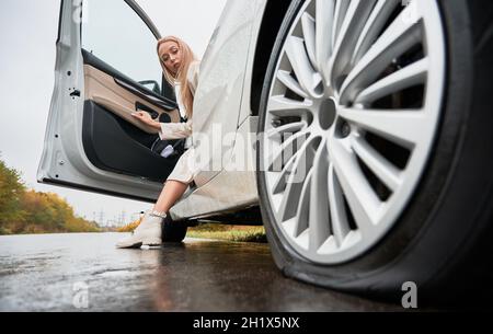 Schöne Frau auf einer Straße stoppte ihr weißes Auto mit durchbrochenem Autoreifen. Die Fahrerin sitzt im Auto und blickt auf das Hinterrad Stockfoto
