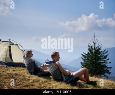 Zwei Wanderer ruhen sich während der Sommerwanderung in den Bergen in Trekkingstühlen auf dem Hügel aus. Angenehme Erholung im Freien in den Bergen. Blauer Himmel mit Wolken und Silhouetten von Hügeln auf dem Hintergrund. Stockfoto