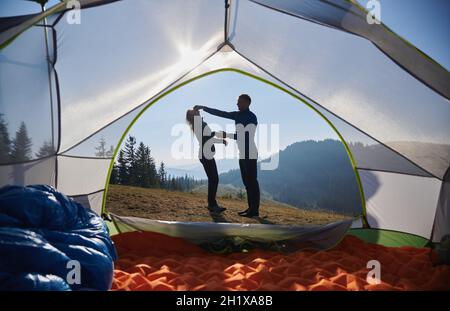 Ein paar Freunde, die Spaß im Freien haben, während sie in den Bergen wandern. Blick vom Innenzelt auf einen Mann, der sanft an der Nase der Freundin zieht und lacht. Lustige Zeit während des Campens in den Bergen. Stockfoto