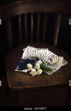 Blumenstrauß aus frühlingshaften weißen und blauen Blumen, eingewickelt in hellgestreiften Stoff auf einem Vintage-Holzstuhl. Dunkler Hintergrund. Draufsicht. Romantisches Geschenk zum Drucken Stockfoto