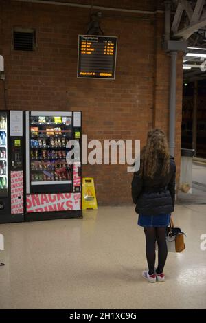 Doncaster, Großbritannien, 22. Mai 2021: Vertikales Bild einer im Winter gekleideten jungen Frau, die das Abfahrtsbrett des Bahnhofs von doncaster liest. Spät in die Nacht Stockfoto