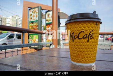 Gelbes Glas aus Papier mit Kaffee für McCafe-Drinks auf dem Tisch gegen die verschwommene Stadt von McDonald's. Hintergrund des Fast-Food-Geschäfts. Fastfood-Res Stockfoto