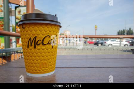 Gelbes Glas aus Papier mit Kaffee für McCafe-Drinks auf dem Tisch gegen die verschwommene Stadt von McDonald's. Hintergrund des Fast-Food-Geschäfts. Fastfood-Res Stockfoto