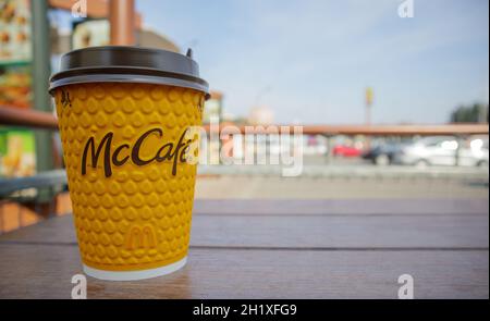 Gelbes Glas aus Papier mit Kaffee für McCafe-Drinks auf dem Tisch gegen die verschwommene Stadt von McDonald's. Hintergrund des Fast-Food-Geschäfts. Fastfood-Res Stockfoto