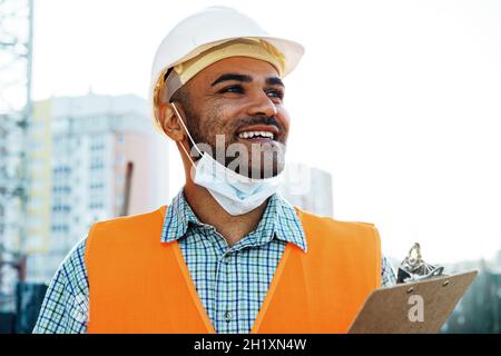 Porträt eines Baumeisters aus gemischter Rasse in Arbeitskleidung und Hardhut mit medizinischer Maske, Nahaufnahme Stockfoto
