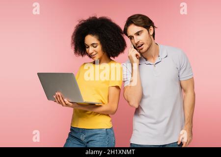 Positive lateinische Frau mit Laptop in der Nähe von Mann sprechen auf Handy isoliert auf rosa Stockfoto