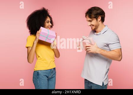 Aufgeregtes multiethnisches Paar lächelt, während es Geschenkschachteln auf rosa isoliert hält Stockfoto
