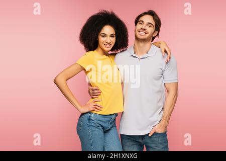Junges und fröhliches interrassisches Paar, das die Kamera anschaut, während es isoliert auf Pink umarmt Stockfoto