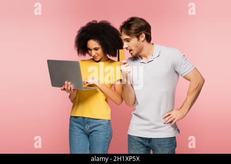 Staunender Mann mit Kreditkarte in der Nähe von hispanischer Freundin mit Laptop isoliert auf rosa Stockfoto