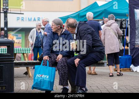 Ein reifes Paar, das auf einer Bank auf einem Straßenmarkt in Lemon Quay in Truro in Cornwall sitzt. Stockfoto