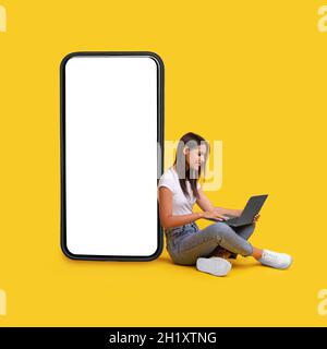 Lächelnde Frau, die einen Laptop benutzt und sich auf einen weißen, leeren Telefonbildschirm stützt Stockfoto