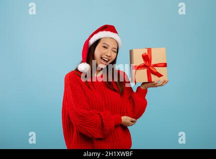 Junge asiatische Dame in Santa Hut hält Geschenkbox und winken, Blick auf die Kamera und lächelnd, blauen Studio-Hintergrund Stockfoto